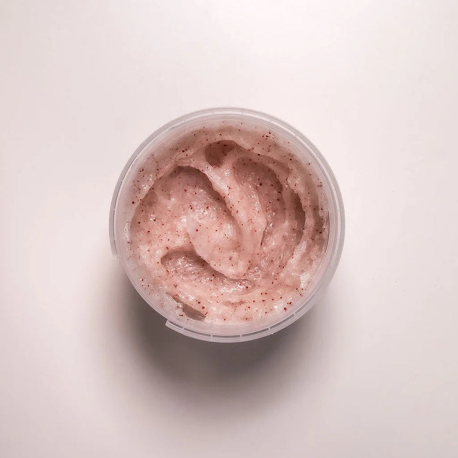 A top-down view of a clear plastic jar filled with a light pink whipped cream or body butter, set against a clean, off-white background.