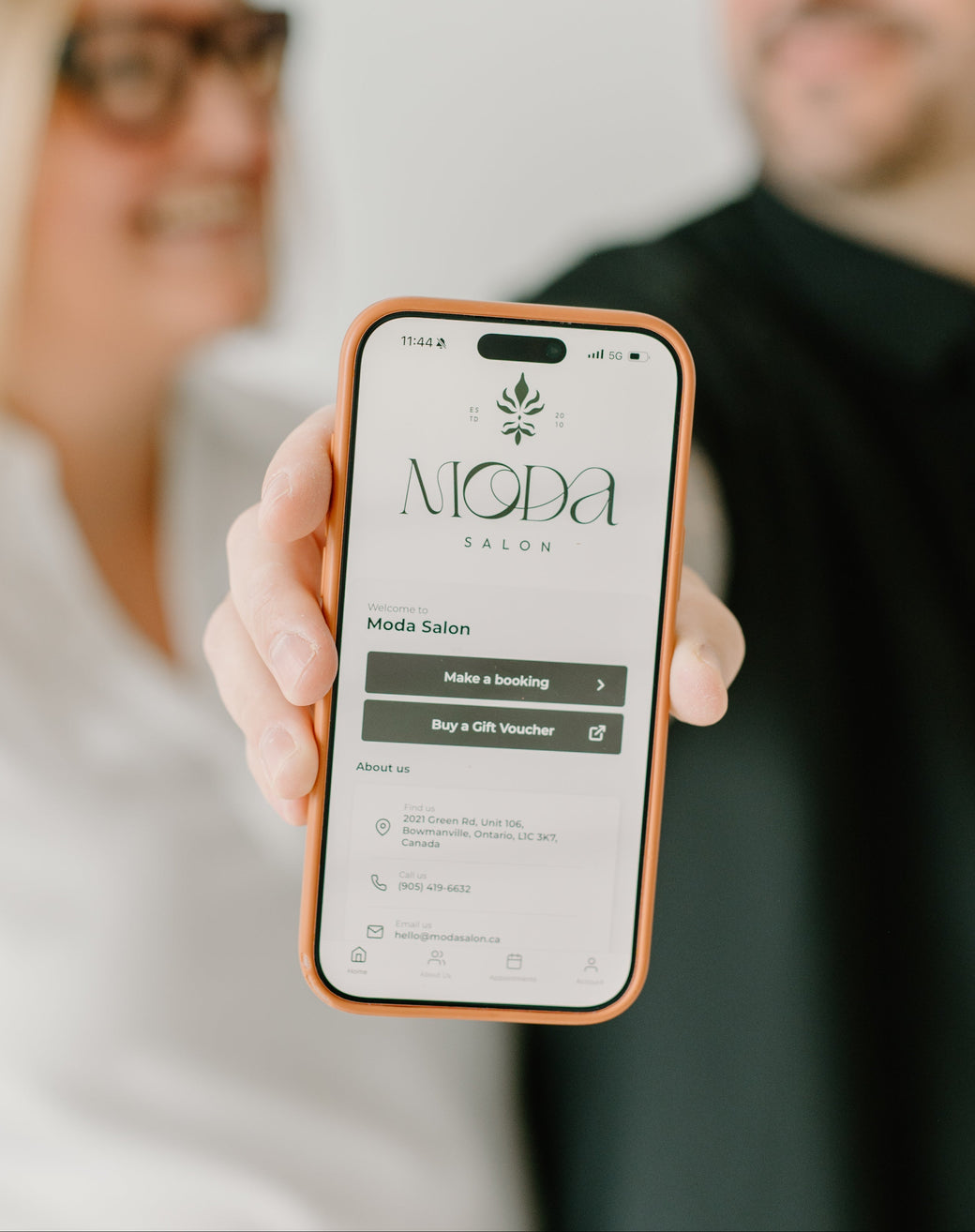Person holding a smartphone displaying a website with two people in the background moda salon phorest app