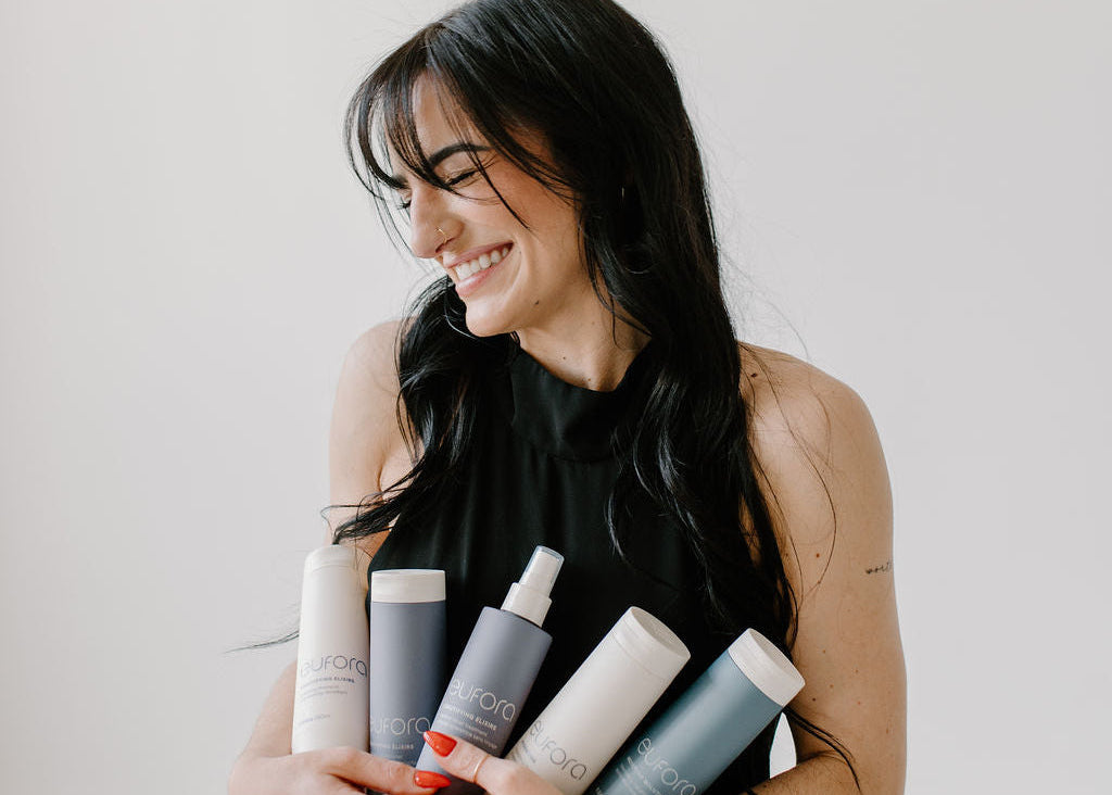 Woman holding multiple hair product bottles against a plain background