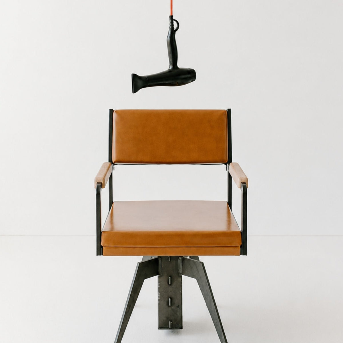 A modern industrial-style chair with tan leather seat and back cushions, black metal frame and legs, and light wood armrests, photographed against a clean white studio background. A black hair dryer hangs from a red cord above the chair.