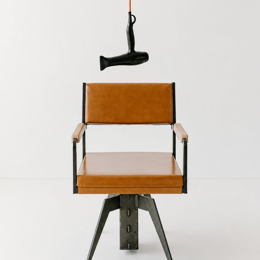 A modern industrial-style chair with tan leather seat and back cushions, black metal frame and legs, and light wood armrests, photographed against a clean white studio background. A black hair dryer hangs from a red cord above the chair.