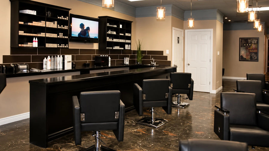 A modern salon interior with a long black reception counter, black styling chairs, wall-mounted shelves, pendant lights, and a television mounted above the counter.