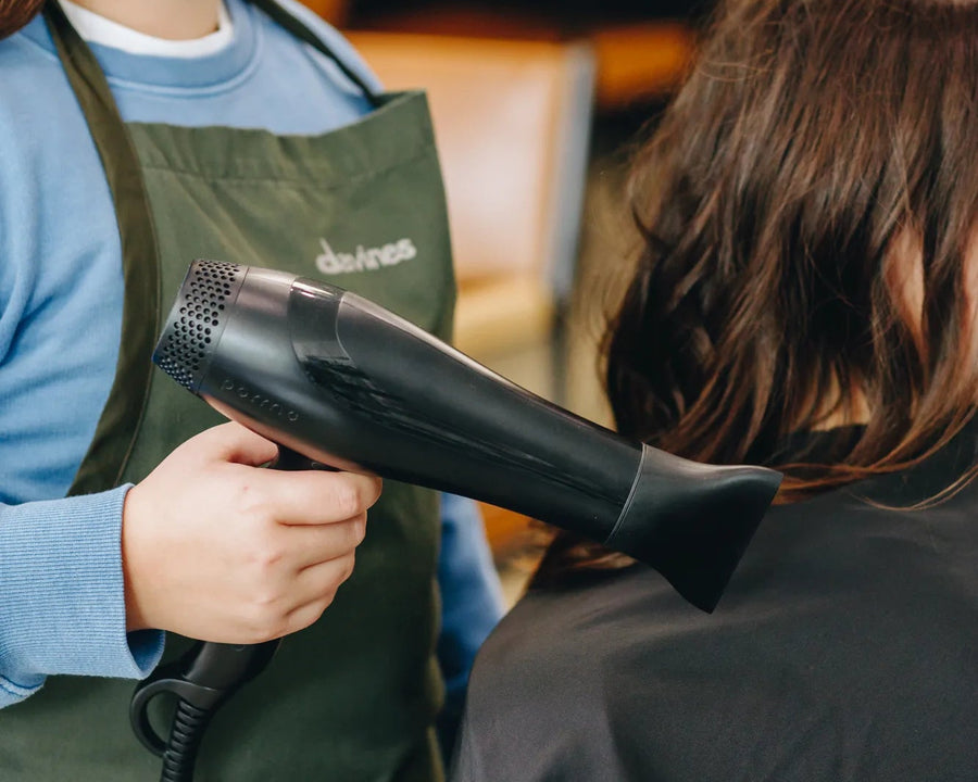 A young girl with long brown hair, wearing a green salon apron over a blue shirt, holds a black hair dryer while styling another person's hair in a salon setting.