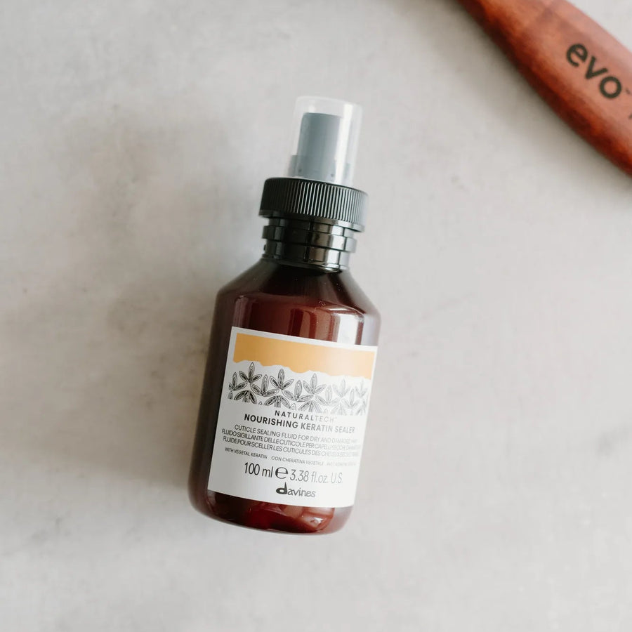 A 100ml bottle of evoCosmetics 'Nourishing Hair Oil' in an amber glass bottle with a black spray cap, styled on a light marble surface next to a wooden hairbrush.