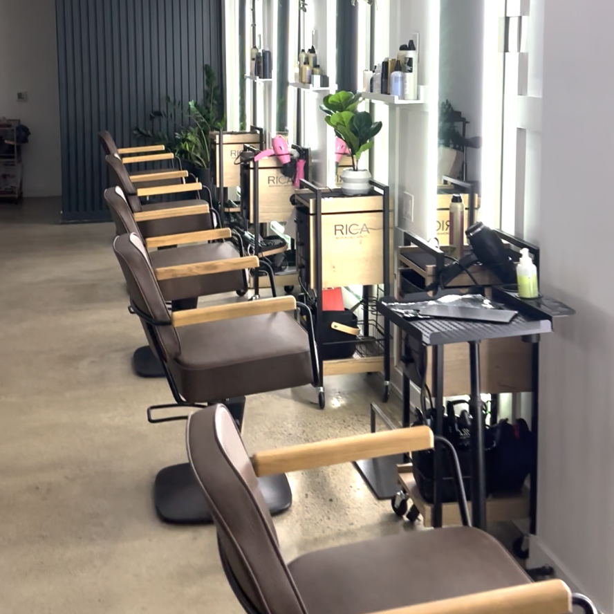 A salon or beauty studio interior with multiple beige and brown chairs arranged along a long row of mirrors, with black styling tables and small decor plants.